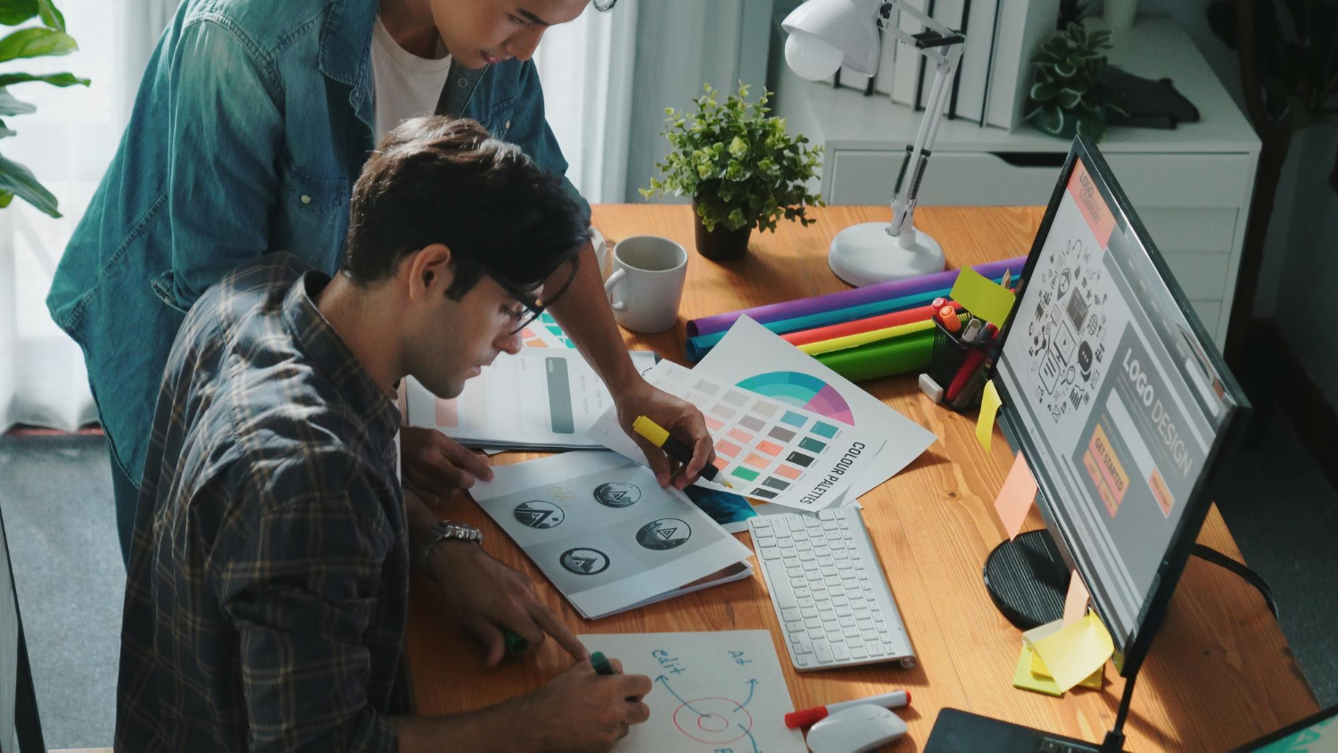Two designers working on logo design concepts with color palettes, sketches, and computer tools, showcasing creativity in modern branding 2025
